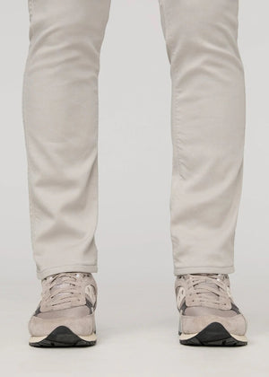 A person stands facing forward on a plain light background, wearing DUER NO SWEAT ATHLETIC TAPER PANTS IN MIST and matching light gray sneakers, feet slightly apart.