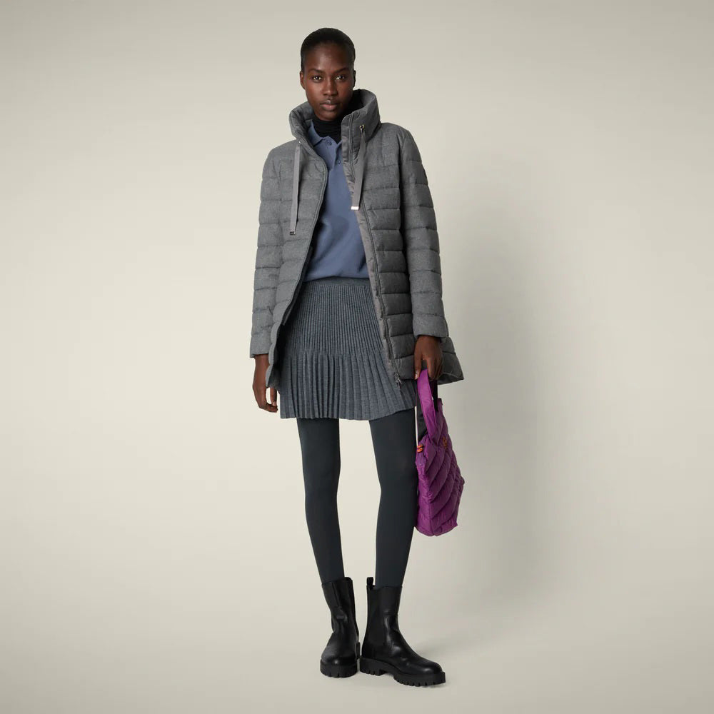 A woman models the SAVE THE DUCK - JUNE PUFFER COAT by SAVE THE DUCK, paired with a blue sweater, pleated grey skirt, black leggings and boots. She holds a purple bag against a plain light background.