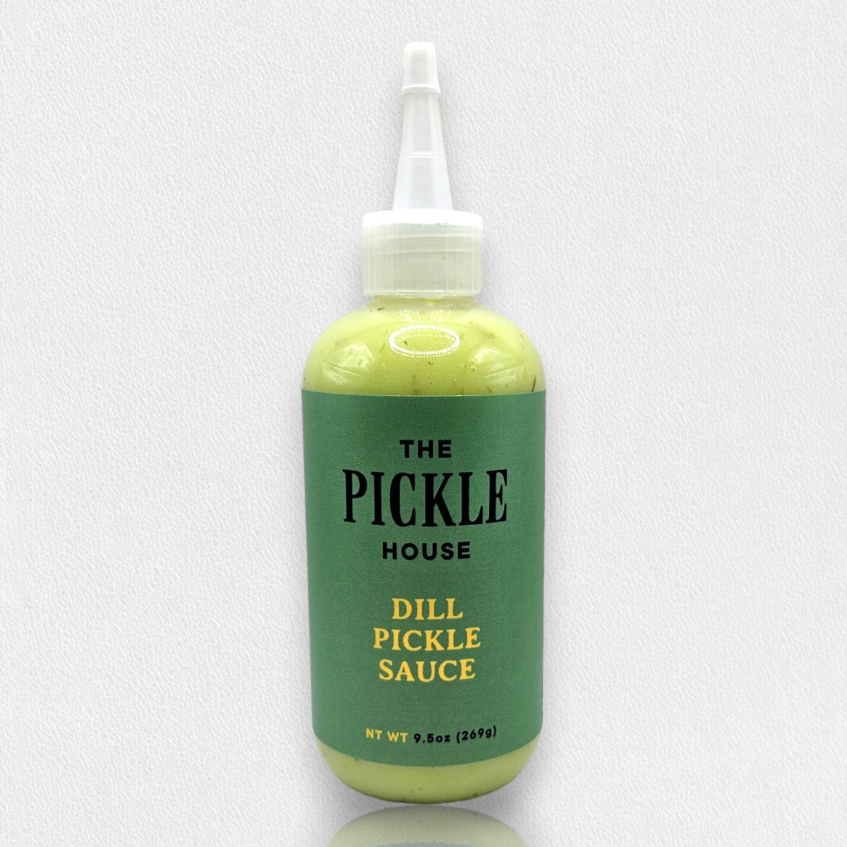 A 9.5-oz squeeze bottle of THE PICKLE HOUSE brand DILL PICKLE SAUCE, with a green label and white cap, sits on a light gray textured background. This Dill Garlic sauce is perfect as a marinade or salad dressing.