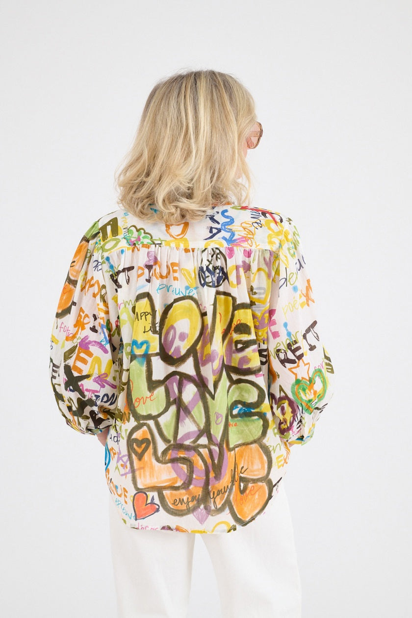 A blonde woman wearing large brown sunglasses and the STORIATIPIC COCO LOVE BLOUSE IN WHITE—a graffiti-patterned cotton top for natural comfort—stands against a white background with her hands loosely clasped.