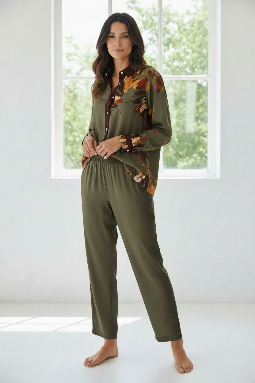 A woman stands barefoot in a sunlit room, her hands casually in her pockets. She wears the FRANCO FERRARI - SILK BLOUSE IN OLIVE, featuring a botanical print, paired with solid olive pajama pants. Behind her, a large window fills the space with natural light.