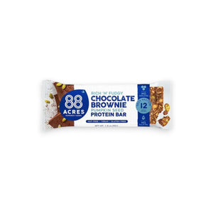 A 88 Acres Dark Chocolate Brownie High Protein Bar wrapper rests on a white wooden surface. The predominantly white and blue wrapper features the brand name "88 Acres" alongside the product name "Rich 'n' Fudgy Dark Chocolate Brownie Pumpkin Seed High Protein Bar." Text highlights that it is nut-free, vegan, and gluten-free.