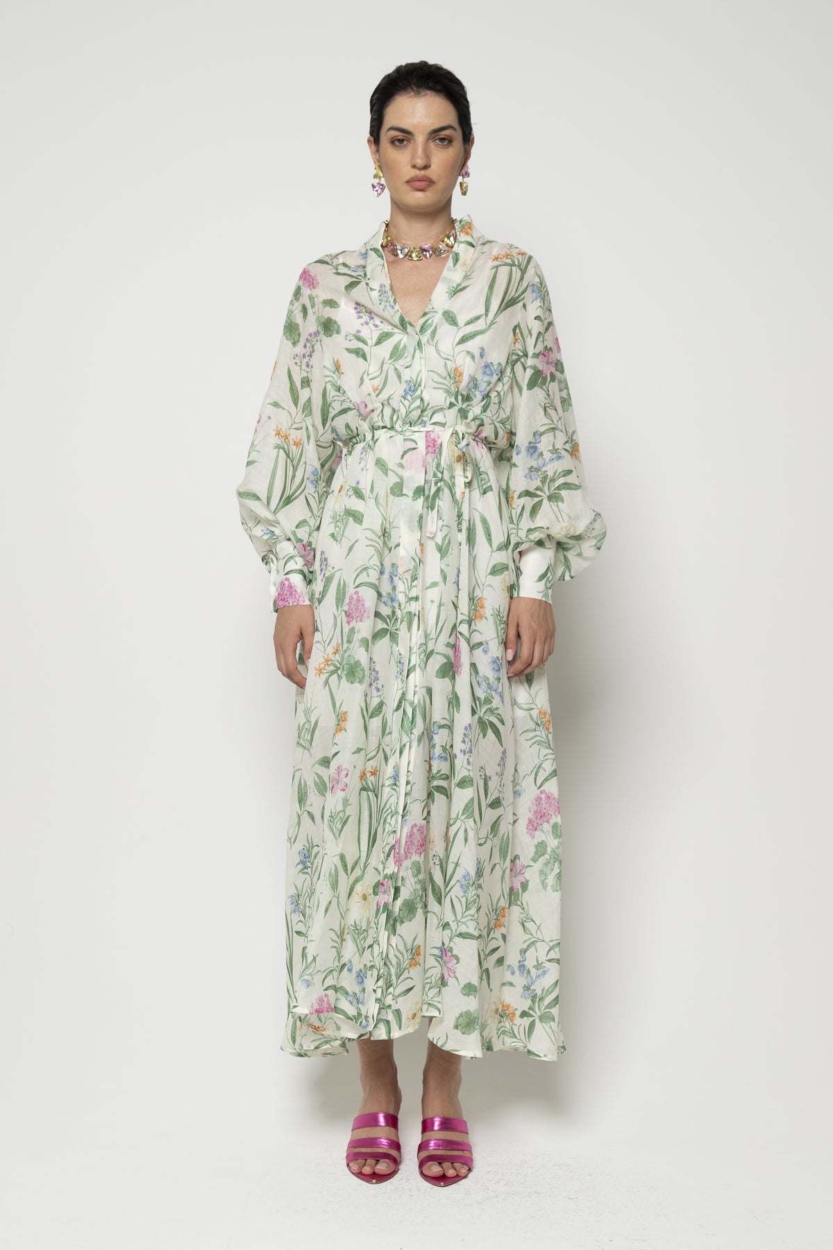 Against a plain background, an individual wears the LAVI COUTURE - ARTEMISIA GREEN FLORAL DRESS, showcasing its luxurious pastel botanical print. They pair it with a colorful necklace and earrings, pink strappy sandals, and neat hair for an elegant look.