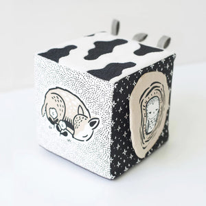 The WEE GALLERY - BABY ANIMALS SOFT BLOCK by WEE GALLERY is a fabric cube adorned with black and white designs that include adorable baby animals, such as a sleeping fox and its cub on one side. This sensory development toy features various textures, including polka dots and stripes, and includes two fabric loops on one edge to aid in tactile exploration.