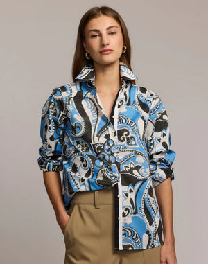 A woman with long brown hair wears the HINSON WU Reese Long Sleeve Shirt in Dancing Paisley, featuring bold blue, black, and white patterns, paired with beige trousers. She poses with a hand in her pocket against a plain background.