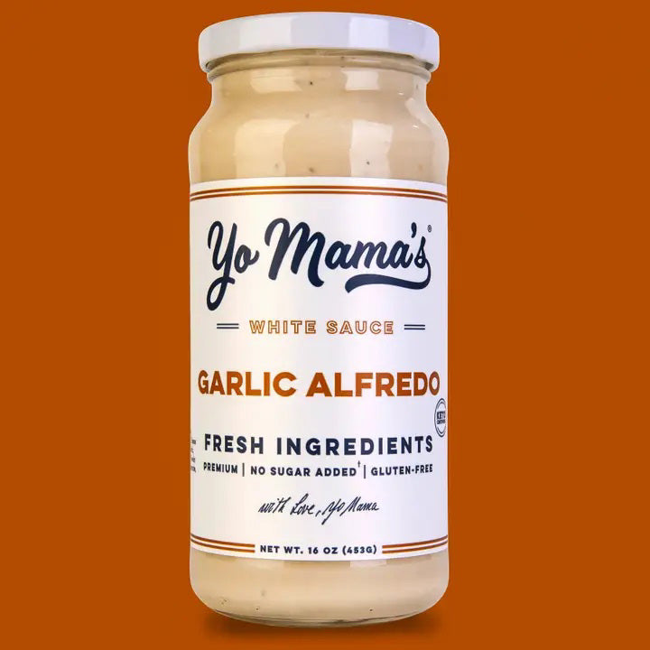 A bowl of green beans topped with creamy YO MAMA'S - ROASTED GARLIC ALFREDO sauce and crispy onions sits on a gray-striped napkin, beside a jar of YO MAMA'S FOODS Garlic Alfredo sauce and white salad servers.