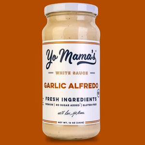 A bowl of green beans topped with creamy YO MAMA'S - ROASTED GARLIC ALFREDO sauce and crispy onions sits on a gray-striped napkin, beside a jar of YO MAMA'S FOODS Garlic Alfredo sauce and white salad servers.