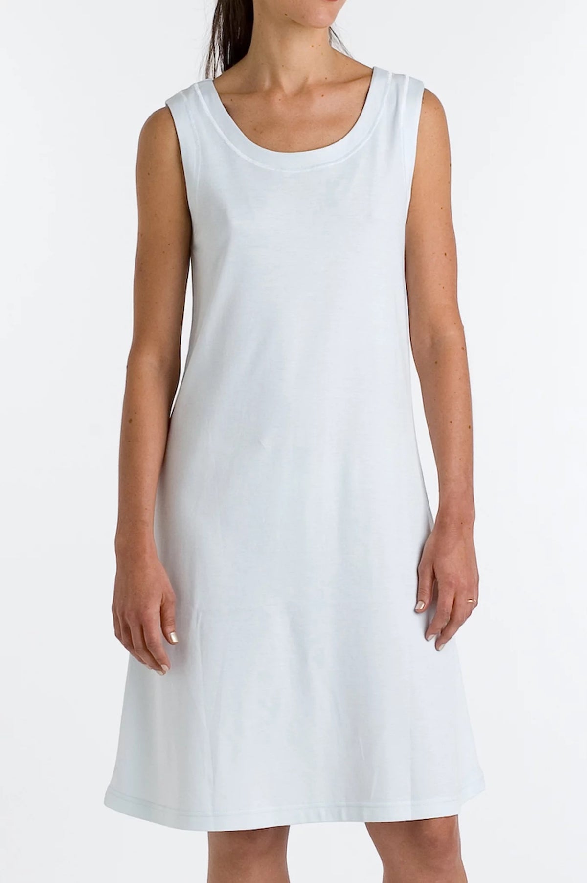 A woman with long hair wearing a simple, sleeveless P. JAMAS - Butterknit Sleeveless Short Gown by FASHIONVIEWS stands against a plain background. The knee-length white dress, made of soft pima cotton knit, features a round neckline and her arms relax by her sides.