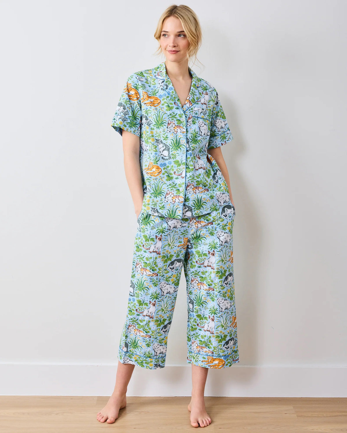 Wearing PRINTFRESH&#39;s &quot;Must Love Cats&quot; cropped pajama set, made of organic cotton poplin, a person stands barefoot against a white wall. The colorful, cat-themed design features short sleeves and cropped pants as they smile slightly.