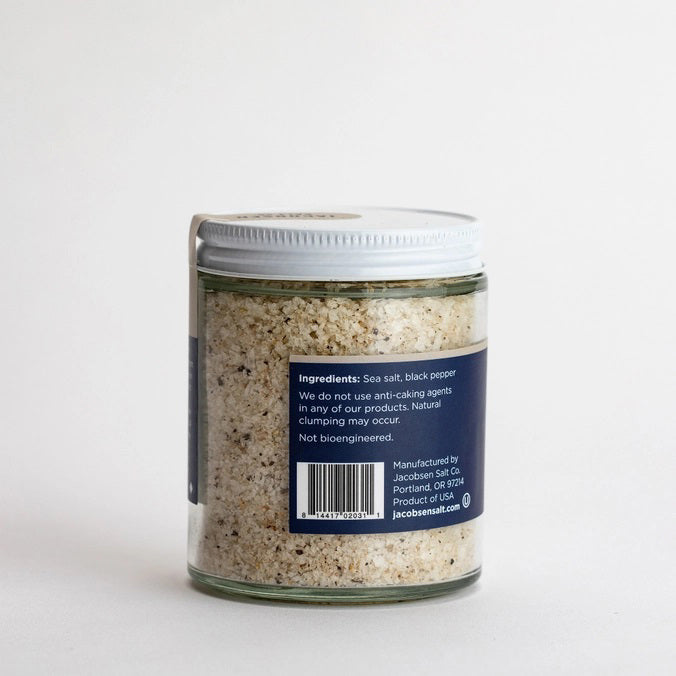 A small glass jar of JACOBSEN SALT CO. JACOBSEN SALT - INFUSED BLACK PEPPER SALT with a white lid rests on a light beige surface against a light gray background. The dark label with white text highlights this floral, spicy blend.