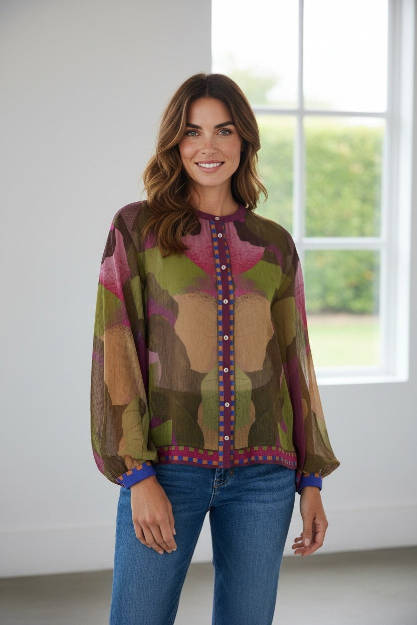 A woman stands indoors, smiling, wearing the FRANCO FERRARI - GERRY SILK TOP featuring an abstract botanical print and button details, paired with blue jeans. Sunlight from a large window illuminates the room, with greenery visible outside.