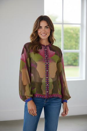 A woman stands indoors, smiling, wearing the FRANCO FERRARI - GERRY SILK TOP featuring an abstract botanical print and button details, paired with blue jeans. Sunlight from a large window illuminates the room, with greenery visible outside.