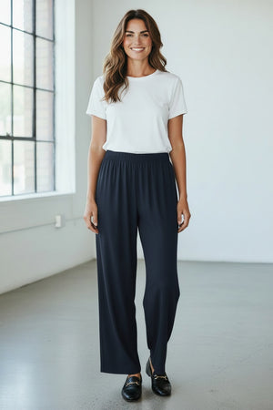A person wearing MEIMEIJ PULL ON STRAIGHT LEG LONG PANTS in black and a red top, shown from the waist down, stands against a light textured background.