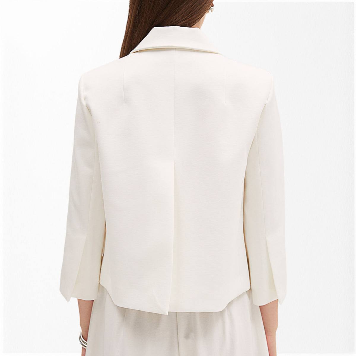A person is wearing the MEIMEIJ - WHITE SHORT JACKET WITH SCALLOPED POCKETS, paired with a matching white top. The jacket, from the brand MEIMEIJ, features a notched collar and three-quarter sleeves. Complementing the outfit, they accessorize with a delicate necklace and a bracelet on their right wrist.