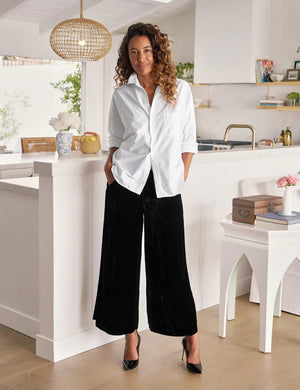 A person indoors wears FRANK & EILEEN's MAISIE Wide Leg Pant in Black Italian Washed Velvet with a white shirt and black pointed-toe heels, standing near a white table topped with books and pink flowers.