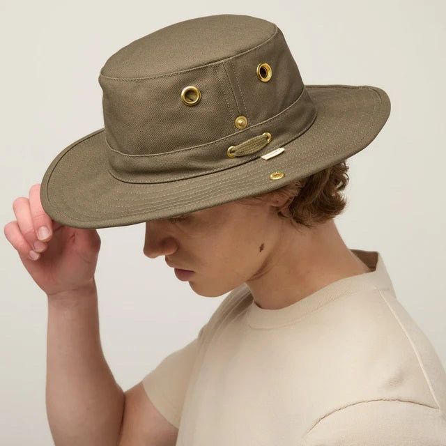 A young person with light skin and wavy brown hair wears the TILLEY - CLASSIC T3 HAT IN OLIVE and a beige crew neck shirt, looking seriously at the camera against a plain light background.