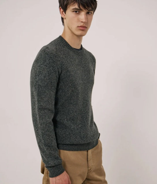A young man with short dark hair wears the HARTFORD Seamless Wool Crew Pullover in Camo Green and tan pants, standing against a plain light-colored background, looking slightly to the side with a neutral expression.
