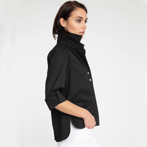 A person with short, dark hair is standing against a plain background, wearing the HINSON WU - AILEEN 3/4 SLEEVE TOP in black with a shirt collar and the sleeves rolled up, paired with white pants. The top features three buttons at the neckline. The individual is looking directly at the camera and smiling slightly.