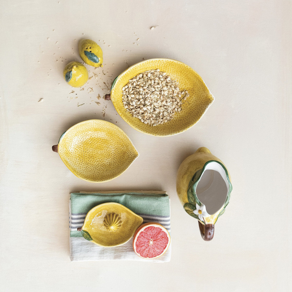A flat lay features CREATIVE COOP’s HAND-PAINTED STONEWARE LEMON SHAPED BOWL, SMALL—one with oats, one with a grapefruit—plus a lemon-shaped pitcher and ceramic lemons atop a folded striped cloth. A delightful citrus-themed display.