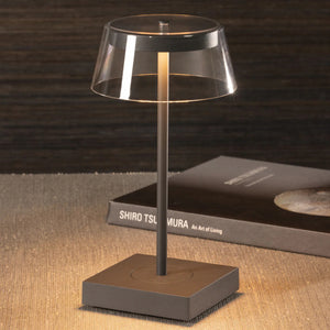 The SARDINIA RECHARGEABLE TABLE LIGHT by ZODAX, with a transparent shade and sleek dark base featuring adjustable brightness, is illuminated on a textured surface beside the book "SHIRO TSUJIMURA: An Art of Living.
