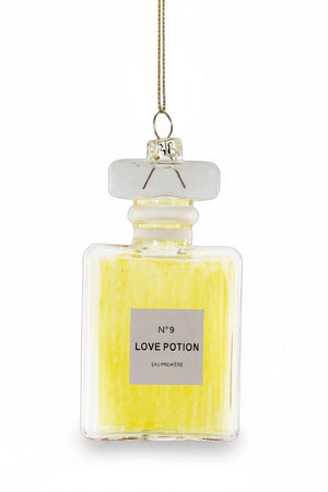 A tall glass bottle labeled "LOVE POTION ORNAMENT" from CODY FOSTER features a decorative stopper and a gold string that enhances its elegant height. Filled with a captivating yellow liquid, it promises enchantment with every glance.
