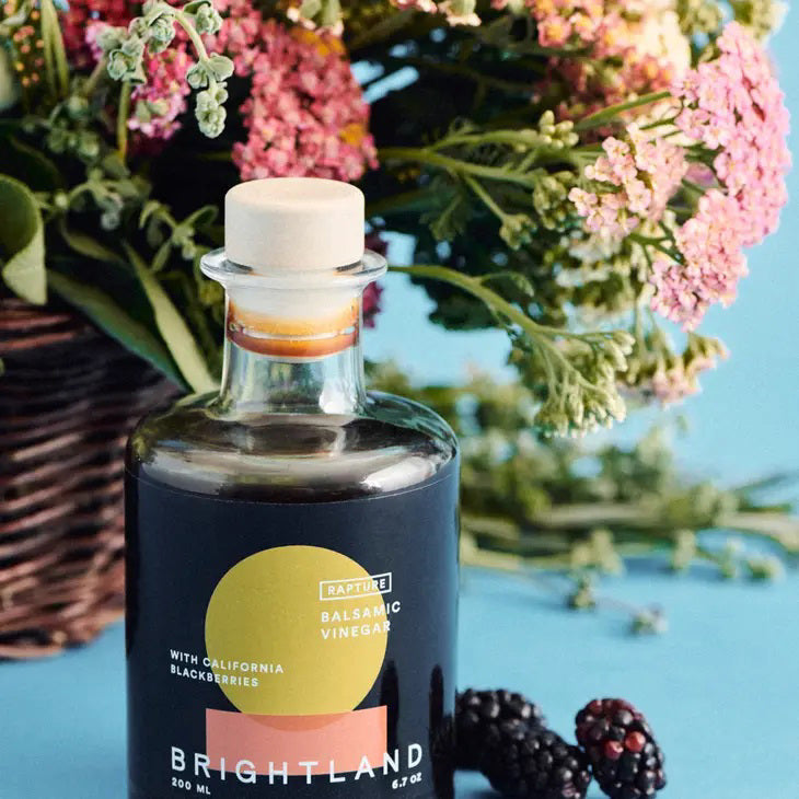 A 200 ml bottle of BRIGHTLAND - &#39;RAPTURE&#39; Blackberry Balsamic Vinegar made with California Zinfandel grapes and triple crown blackberries. The bottle has a round body, a short neck, and is sealed with a light-colored cork stopper. Its label boasts a modern design featuring a yellow and orange circle against a black background.