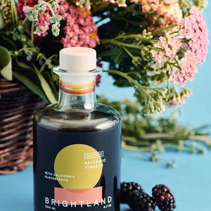 A 200 ml bottle of BRIGHTLAND - 'RAPTURE' Blackberry Balsamic Vinegar made with California Zinfandel grapes and triple crown blackberries. The bottle has a round body, a short neck, and is sealed with a light-colored cork stopper. Its label boasts a modern design featuring a yellow and orange circle against a black background.