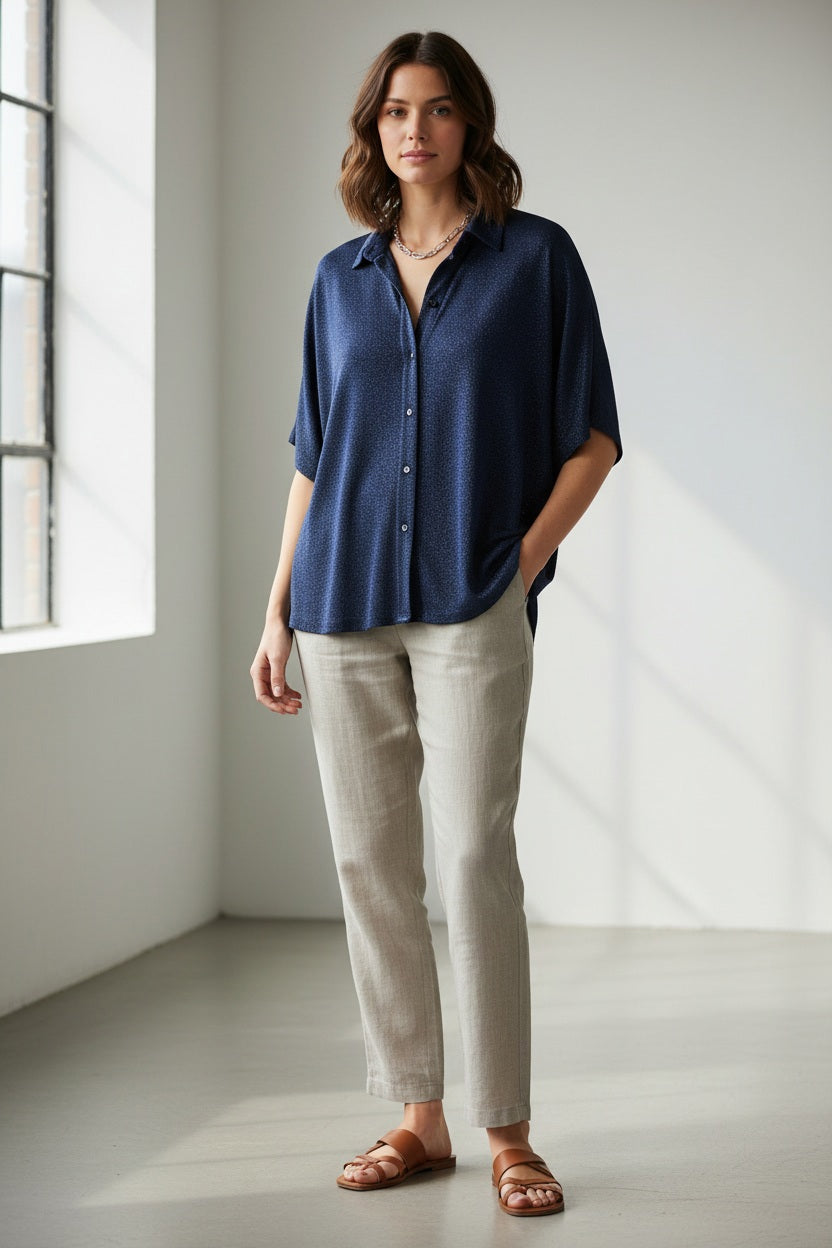 A person in a MOMONI - BROOKLYN blue shirt with a classic collar, paired with colorful beaded necklaces and tucked into striped pants. The loose-fitting, short-sleeve button-up stands out against the plain light background.