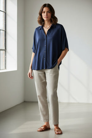 A person in a MOMONI - BROOKLYN blue shirt with a classic collar, paired with colorful beaded necklaces and tucked into striped pants. The loose-fitting, short-sleeve button-up stands out against the plain light background.