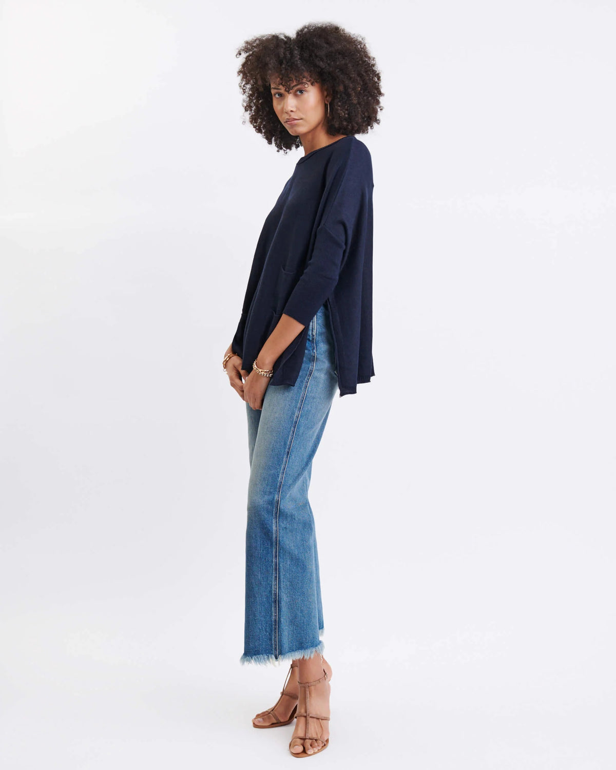 A woman stands in front of a gray wall and a green potted plant, wearing the MERSEA - CATALINA CREWNECK SWEATER IN NAVY by MERSEA with an oversized fit, paired with white pants, layered necklaces, and a bracelet, as she looks slightly to the side.