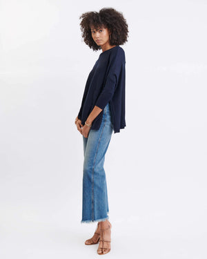 A woman stands in front of a gray wall and a green potted plant, wearing the MERSEA - CATALINA CREWNECK SWEATER IN NAVY by MERSEA with an oversized fit, paired with white pants, layered necklaces, and a bracelet, as she looks slightly to the side.