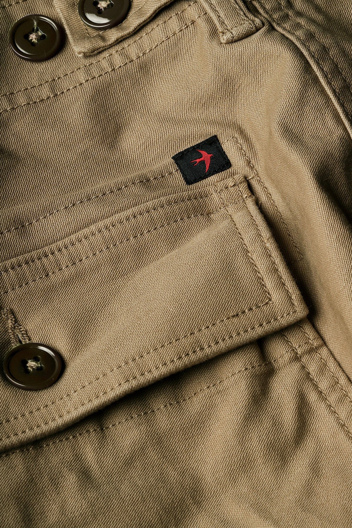 The RELWEN Supply Pant in Worker Khaki is a pair of light brown chinos inspired by classic military utility pant design, featuring side adjuster tabs, a button fly, broken twill fabric, and cuffs at the ankles with a subtly tapered leg silhouette—laid flat on a white background.