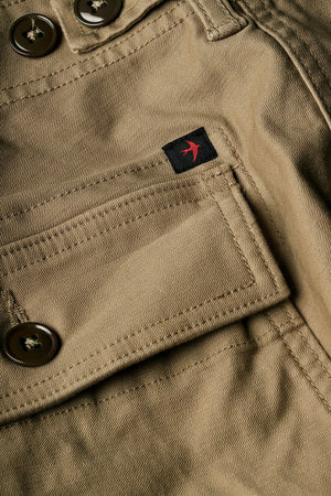 The RELWEN Supply Pant in Worker Khaki is a pair of light brown chinos inspired by classic military utility pant design, featuring side adjuster tabs, a button fly, broken twill fabric, and cuffs at the ankles with a subtly tapered leg silhouette—laid flat on a white background.