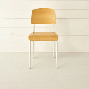 A simple wooden chair with a curved backrest and white metal legs sits on the CHILEWICH MINI BASKETWEAVE RUNNER 2.5X8, made from phthalate-free TerraStrand yarns, against a white wood-paneled wall.