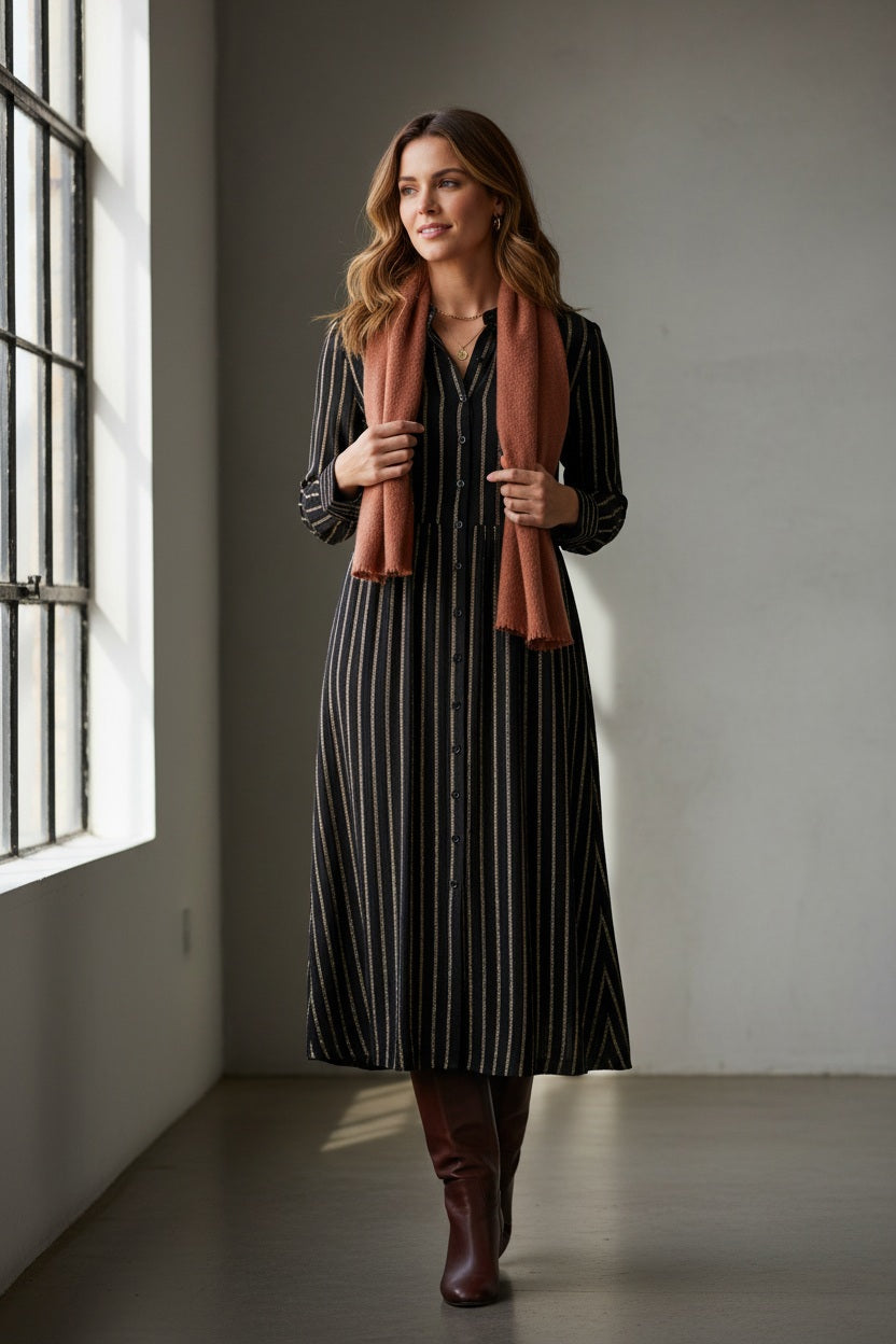 A woman in the MOMONI Aubert Dress in Lurex Gauze stands by a woven chair, gazing down as her hair partially covers her face.