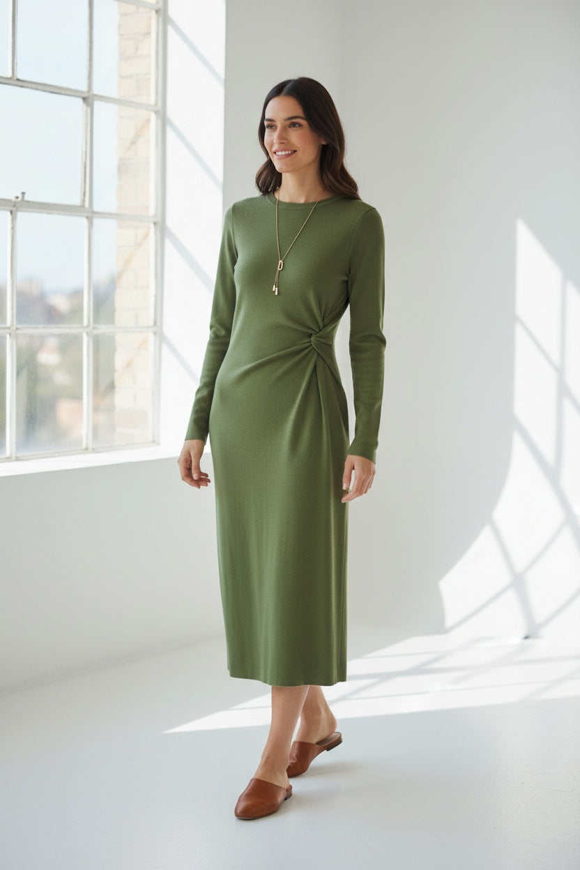 A woman wearing the MEIMEIJ Side Gathered Dress in Palm Green, featuring long sleeves and a midi length with a side knot detail, styled with brown flats and a long necklace, stands in a bright, minimalist room with large windows.