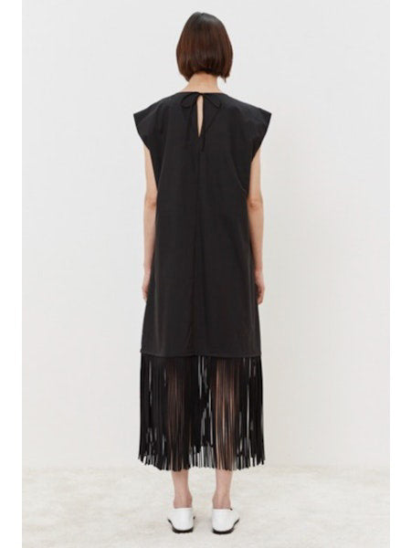 The person models the MEIMEIJ - FRINGE DRESS, a cotton, sleeveless black dress with a V-neckline and knee-length flowing fringes. They pair it elegantly with white shoes against a plain white background.