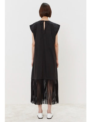 The person models the MEIMEIJ - FRINGE DRESS, a cotton, sleeveless black dress with a V-neckline and knee-length flowing fringes. They pair it elegantly with white shoes against a plain white background.