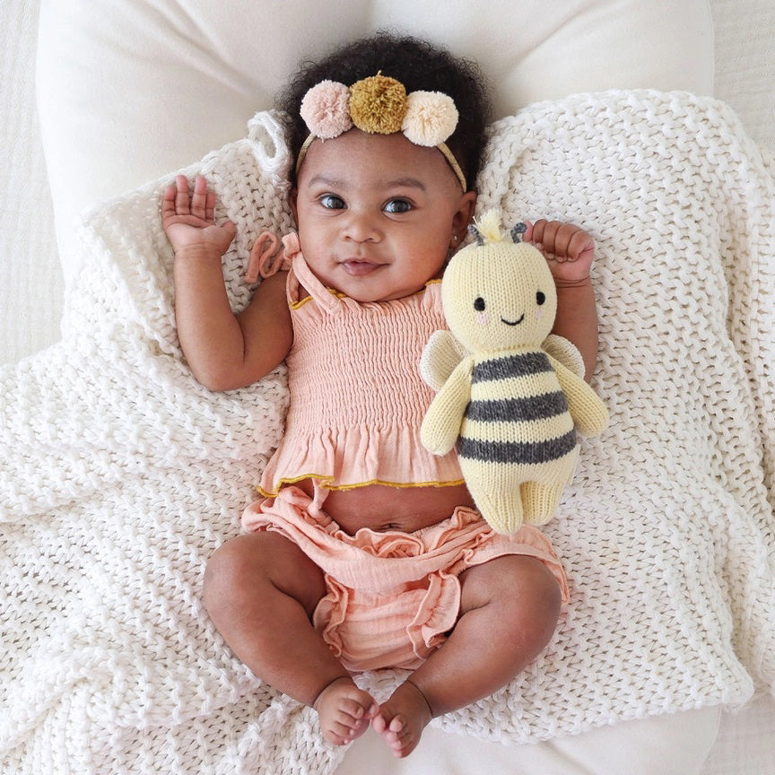 A baby lies on a cozy white blanket, raising both arms. The baby is dressed in a peach outfit and a headband with three pom-poms. Next to the baby is the CUDDLE + KIND - HANDMADE BABY BEE by CUDDLE &amp; KIND, a fair trade, hand-knit toy with black and white stripes—a perfect addition to any collection of baby shower gifts.
