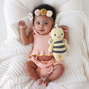 A baby lies on a cozy white blanket, raising both arms. The baby is dressed in a peach outfit and a headband with three pom-poms. Next to the baby is the CUDDLE + KIND - HANDMADE BABY BEE by CUDDLE & KIND, a fair trade, hand-knit toy with black and white stripes—a perfect addition to any collection of baby shower gifts.