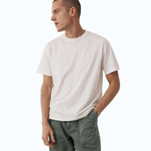 Wearing the HARTFORD - SLUB COTTON CREWNECK T-SHIRT IN OFF WHITE, a short-haired person in green pants stands against a light background with hands loosely clasped, small wrist tattoos visible.