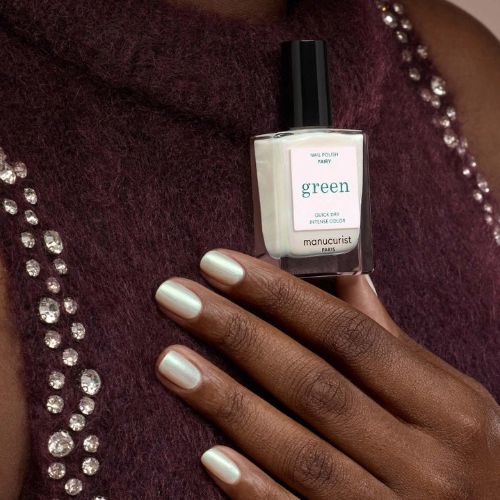 A person with light green painted nails holds a bottle of MANUCURIST INC’s GREEN™ Nail Polish in Fairy, wearing a purple knit top adorned with rhinestone embellishments.