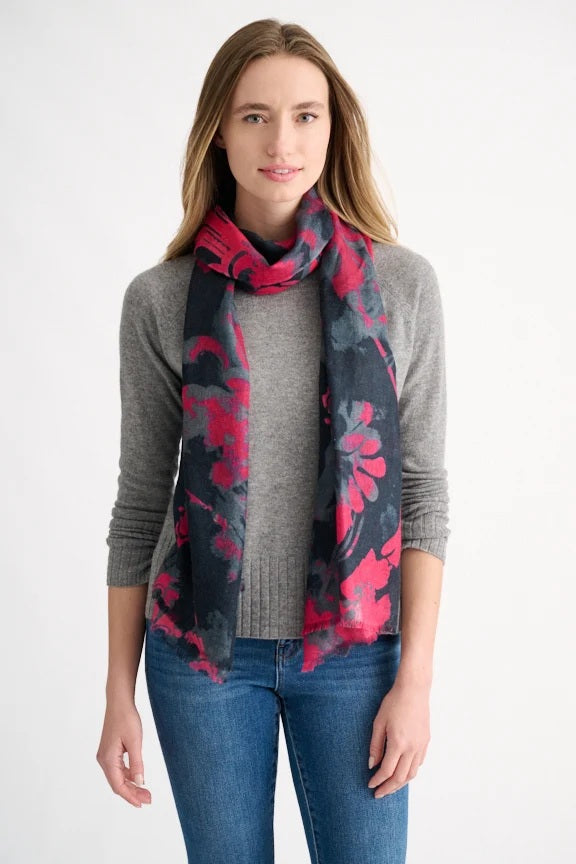 A woman with straight, light brown hair, dressed in a gray sweater and blue jeans, stands facing forward against a plain white background. She accessorizes her outfit with the KINROSS TAPESTRY SCARF IN CRANBERRY by KINROSS—a lightweight, elegant scarf featuring rich red and dark floral patterns.