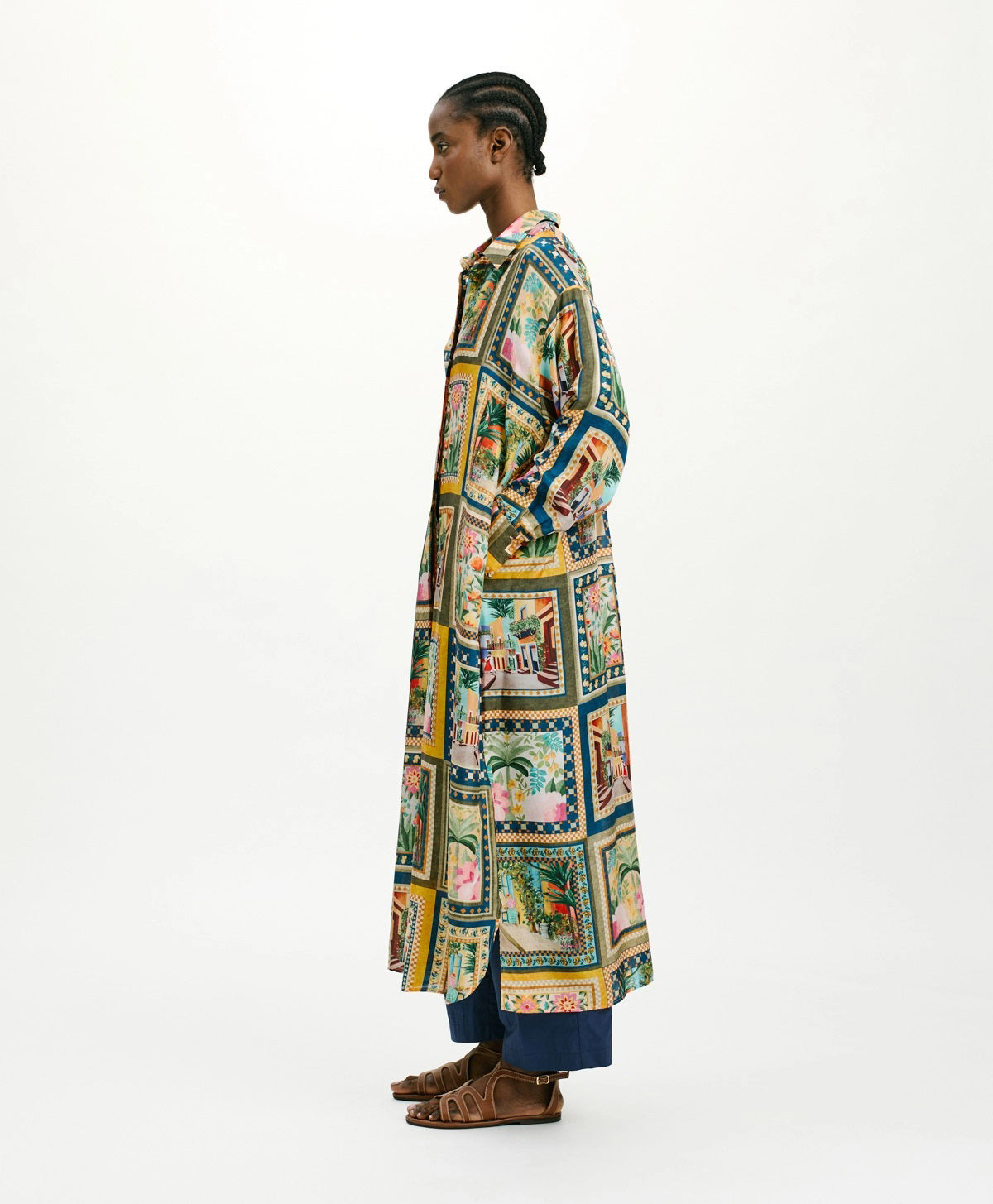 A person wears the MOMONI JAU DRESS, a colorful, artistic printed shirt dress, over blue high-waisted trousers against a plain backdrop. Brown sandals complete the effortless style as they gaze sideways.