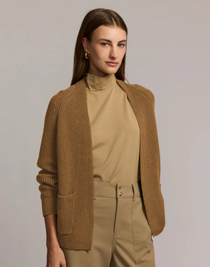 A woman with straight brown hair, in a tan turtleneck and high-waisted tan pants with gold buttons, wears the HINSON WU ANGIE Long Sleeve Cardigan in dark olive green, standing arms crossed against a plain light gray background.
