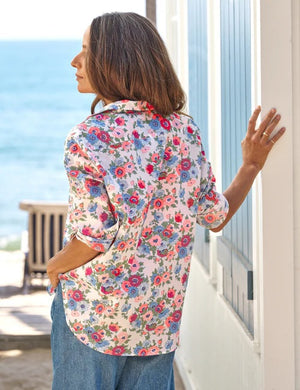 A woman with long brown hair stands smiling in front of a white wall with blue shutters, wearing the FRANK & EILEEN EILEEN Relaxed Button Up in Liberty Fabric—pink and red with blue flowers—paired with blue jeans.