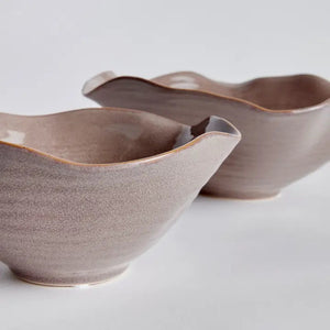 Displayed on a white background are two NAPA HOME AND GARDEN RIVO decorative bowls in beige with wavy, scalloped edges and glossy plum glaze, showcasing natural ceramic artistry. The bowls vary in size, with one being slightly larger.