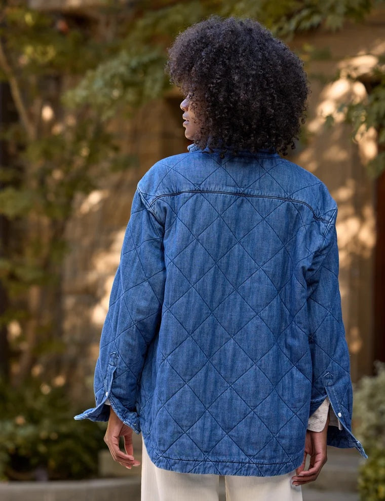 A person holds open a blue FRANK &amp; EILEEN - EILEEN Quilted Shirt Jacket in Famous Denim Distressed Indigo Wash, revealing its interior pocket and distinctive diamond quilted pattern. The person is wearing a cream-colored, ribbed top, with a blurred background.