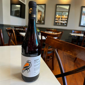 A bottle of TSIAKKAS ‘MOUKLOS’ MAVRO 2024 red wine with a label featuring an illustration of an orange and black bird, sitting on a white table in a restaurant.