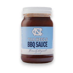 A jar of Cackalacky - Carolina Tar Heels BBQ Sauce is placed on a wooden surface, alongside a light blue football helmet and football. The display is artistically enhanced with scattered blue and white paper shreds, highlighting the vibrant appeal of this savory tomato-based barbecue sauce from Cackalacky.